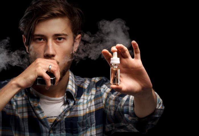 Man is vaping and holding e-liquid. Black background.