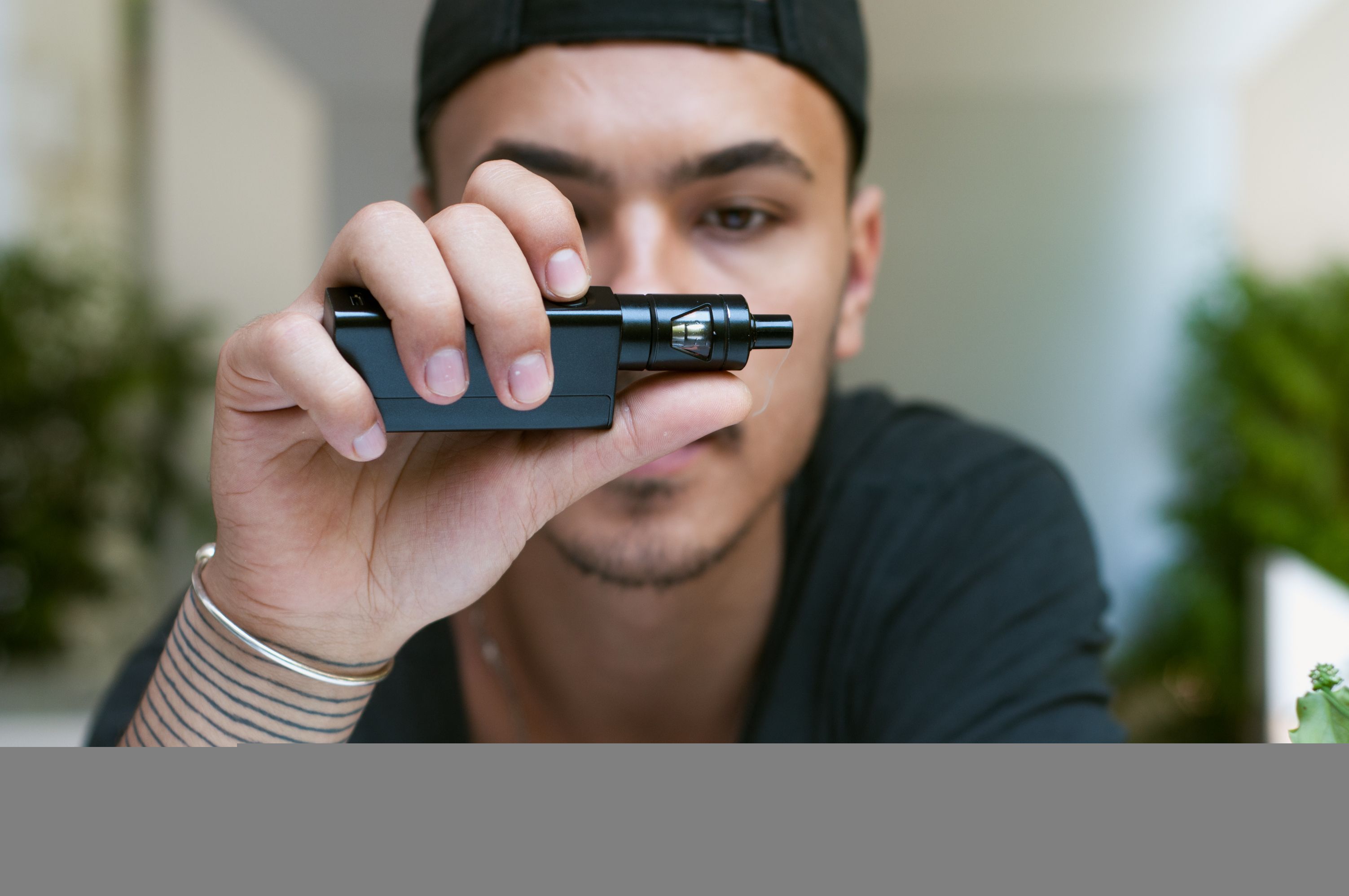 young bearded man holds an electronic cigarette (e-cig) and exhaling a cloud of vapor. guy vaping in sunglasses with tattoos