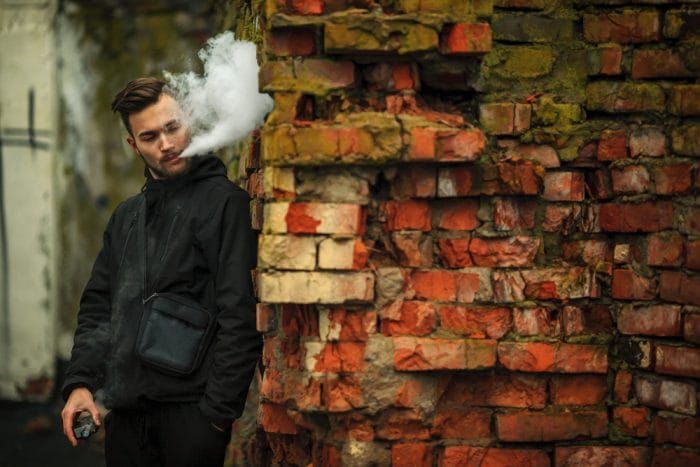 Vape man. Photo of a handsome young white guy with modern haircut in the black clothes vaping and letting off steam from an electronic cigarette near