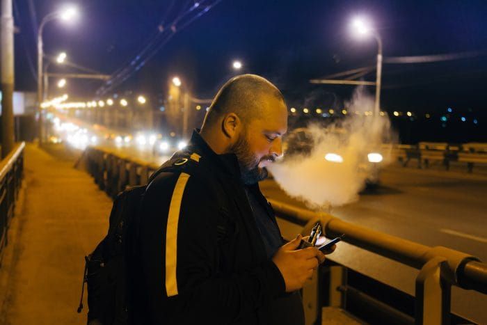 Vaping by electronic cigarette fat man using smart-phone on the bridge at night city.