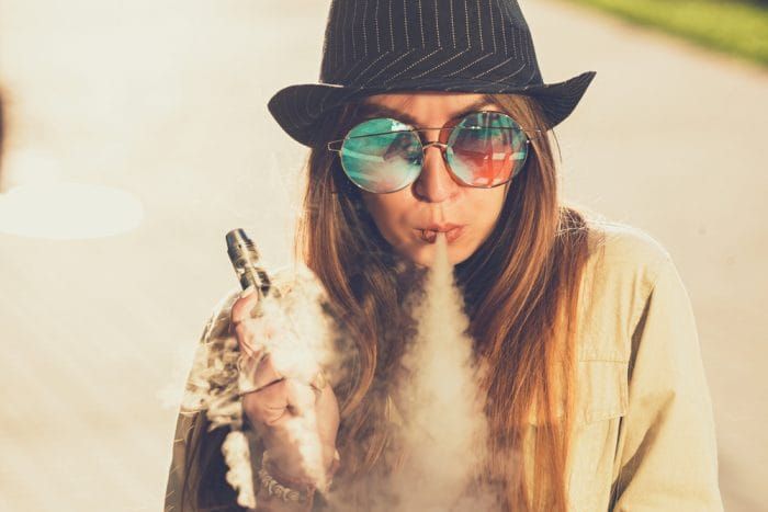 Pretty young hipster woman in black hat vape ecig, vaping device at the sunset. Toned image