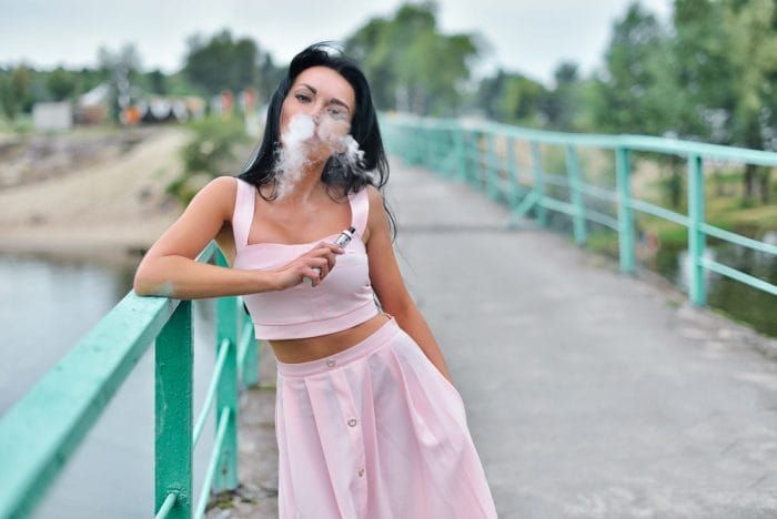 Vaping. Young beautiful woman smoking ( vaping ) e-cigarette with smoke outdoors. Vapor concept.