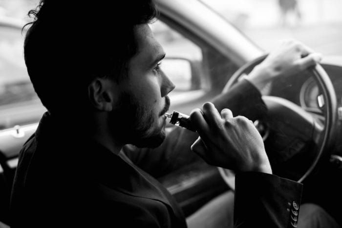 View from the side of a young man smoking an e-cigarette as he drives his car on an urban street