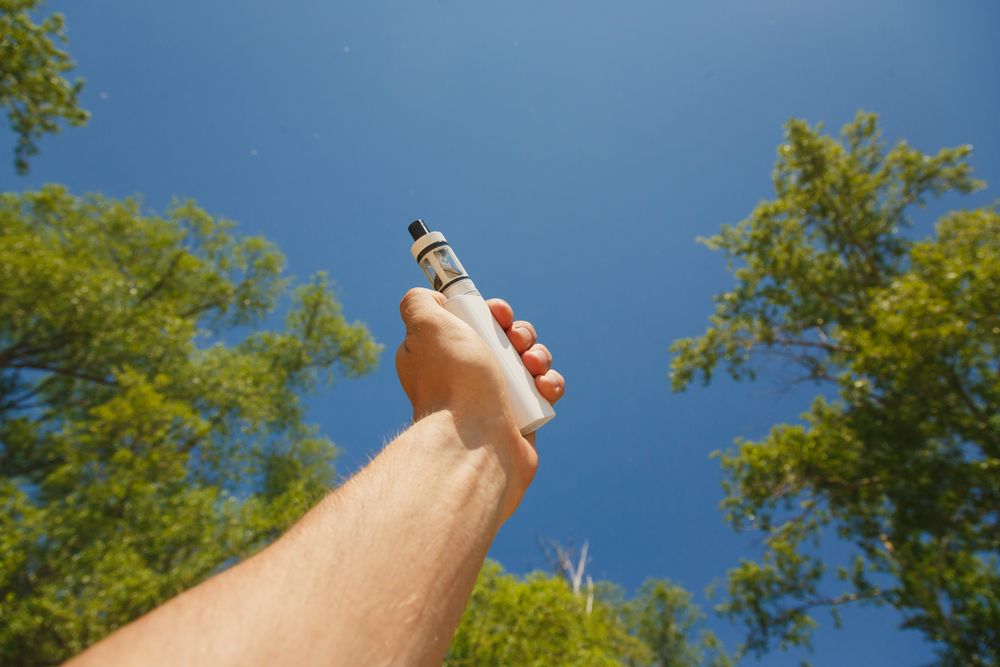 Close up of a man hand holding an electronic cigarette on sky background. Vape in summer, eco-friendly