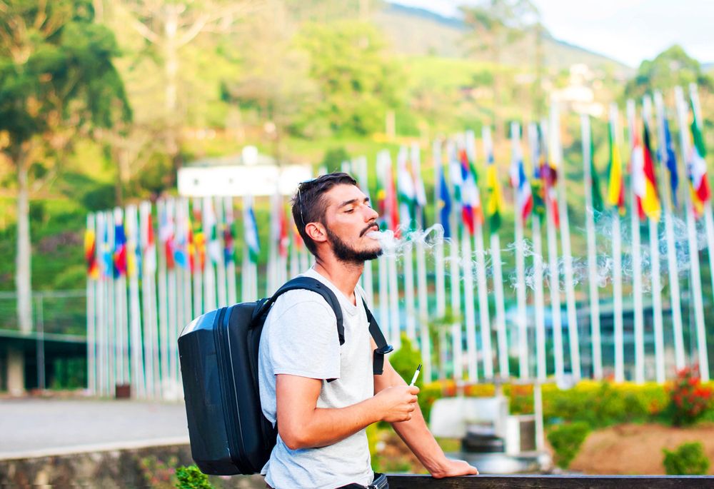 A handsome guy with a beard smokes an electronic cigarette on the background of mountains and flags of different countries